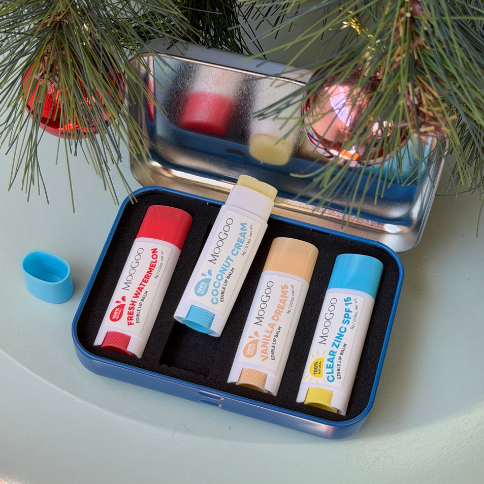 A set of four MooGoo lip balm tubes arranged in a blue metal case on a light surface, with pine branches and holiday ornaments in the background. The balm tubes are in different colors and flavours: red fresh watermelon, light blue coconut cream, beige vanilla dream, and dark blue clear zinc spf15.