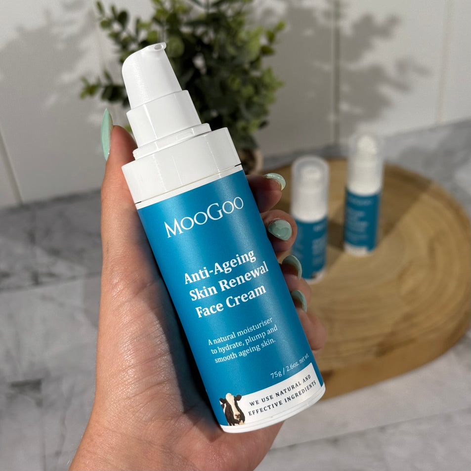 MooGoo Anti-Ageing Skin Renewal Face Cream in a blue bottle, held by a hand with green nails, with a plant and other MooGoo products in the background on a wooden board and tiles.