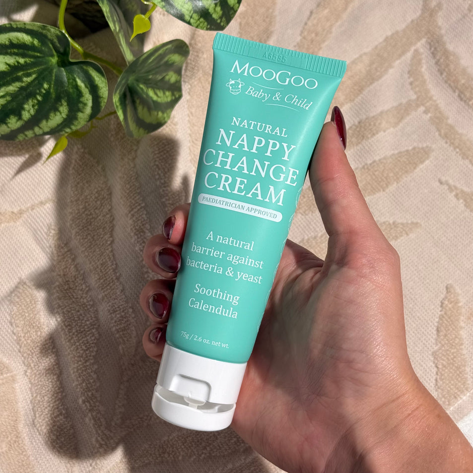 Hand holding a tube of MooGoo Baby & Child Natural Nappy Change Cream on a beige towel background with a green plant, paediatrician approved, calms and protects baby's skin against nappy rash.