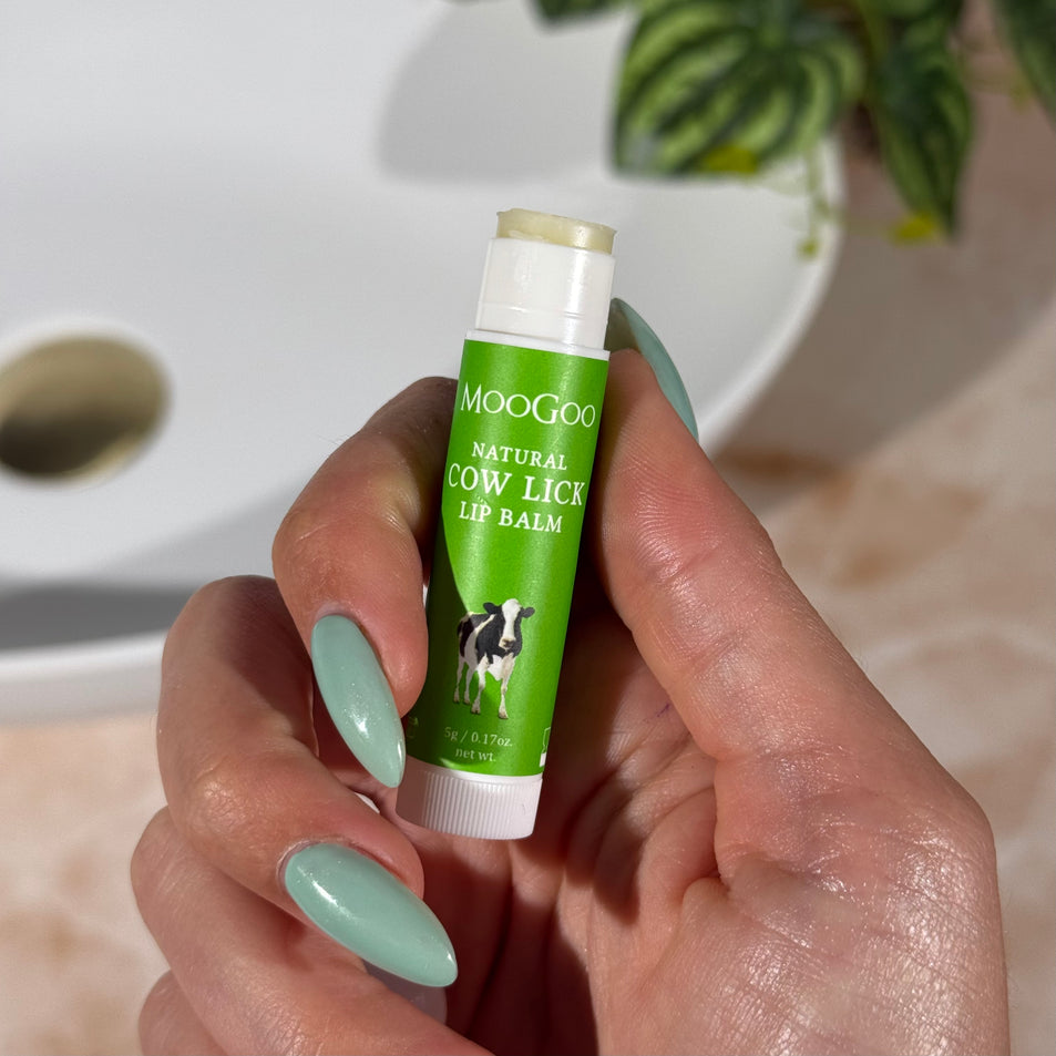 Hand holding a green tube of MooGoo Natural Cow Lick Lip Balm with a plant and bathroom sink in the background.