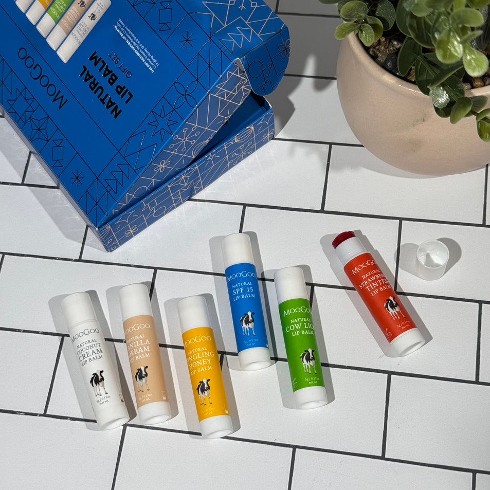 MooGoo Natural Lip Balm Christmas Gift Set in blue themed packaging on a tiled background, with the box open, displaying six MooGoo Lip Balm flavours in various colours and  a plant in the background.