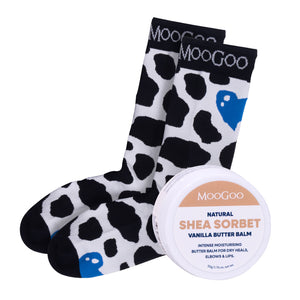 A pair of MooGoo branded socks with a black and white cow print and blue hearts, shown next to a round tin of MooGoo Natural Shea Sorbet Vanilla Butter Balm on a white background. Hydrating foot treatment for dry heels and overnight repair. 