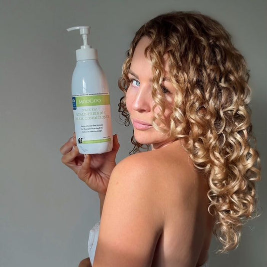 MooGoo Sensitive Scalp Cream Conditioner bottle held by woman with curly blonde hair, nourishing silicone free conditioner for scalp and hair care