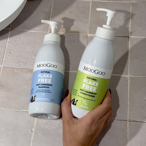 Two MooGoo flake free shampoo bottles, one with a blue label and one with a green label, held by a hand against a tiled bathroom background. Silicone free formula with jojoba and coconut oil to restore dry scalp. 