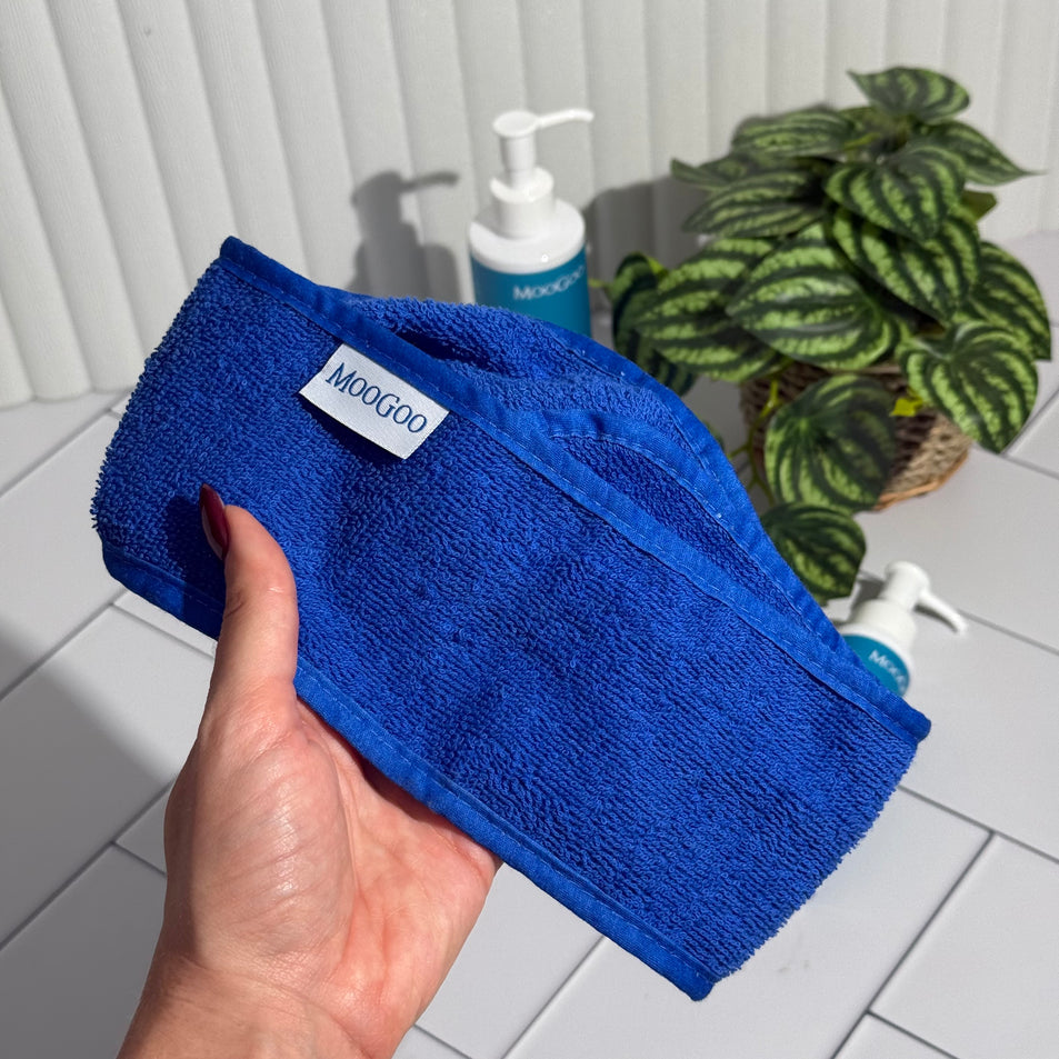 Blue terry toweling Headband with a MooGoo label, held by a hand with MooGoo Face Cleansers and a plant in the background.