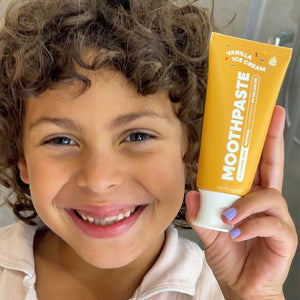 A smiling child with curly brown hair holds up a tube of Moothpaste Vanilla Ice Cream toothpaste. The tube is bright yellow with white text and a white cap, while the child is wearing a light beige top and has light purple painted fingernails.