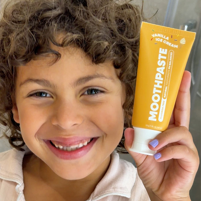 A smiling child with curly brown hair holds up a tube of Moothpaste Vanilla Ice Cream toothpaste. The tube is bright yellow with white text and a white cap, while the child is wearing a light beige top and has light purple painted fingernails.
