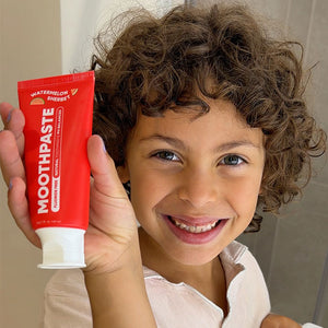 A smiling child with curly brown hair holds up a red tube of Watermelon Kids Moothpaste toothpaste in front of their face. 