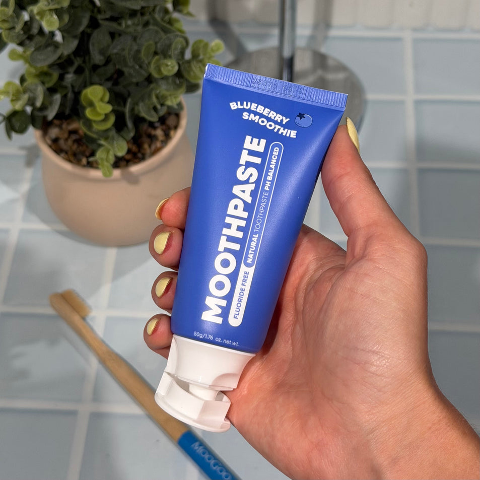 Hand holding MooGoo Mini Moo Kids Blueberry Smoothie flavoured toothpaste, with a toothbrush greenery and blue tiles in the background. Healthy habit building, tastes like blueberries. Fluoride-free formula, safe for kids. Safe if Swallowed. 