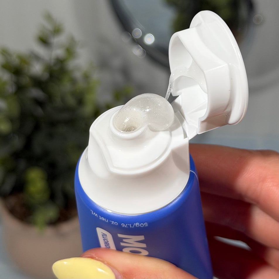 Hand holding a tube of MooGoo Mini Moo Blueberry Smoothie kids toothpaste and squeezing out the clear toothpaste. Healthy habit building, tastes like blueberries. Fluoride-free formula, safe for kids. Safe if swallowed. 