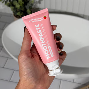 Hand holding MooGoo Mini Moo Kids Strawberries and Cream flavoured toothpaste, with greenery and white tiles in the background. Healthy habit building, tastes like strawberries. Fluoride-free formula, safe for kids. Safe if swallowed.