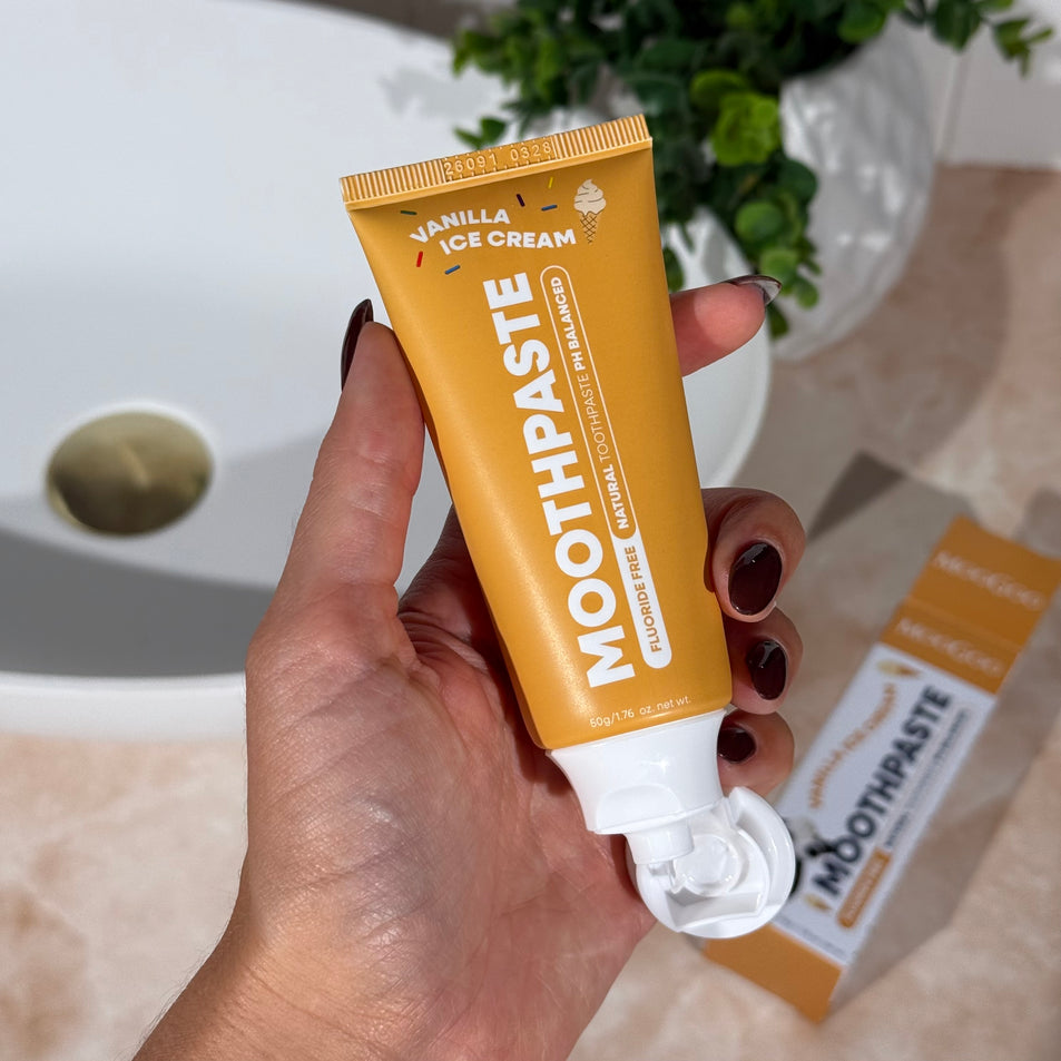 Hand holding MooGoo Mini Moo Kids Vanilla Ice Cream flavoured toothpaste, with greenery and neutral tiles in the background. Healthy habit building, tastes like vanilla. Fluoride-free formula, safe for kids. Safe if swallowed.