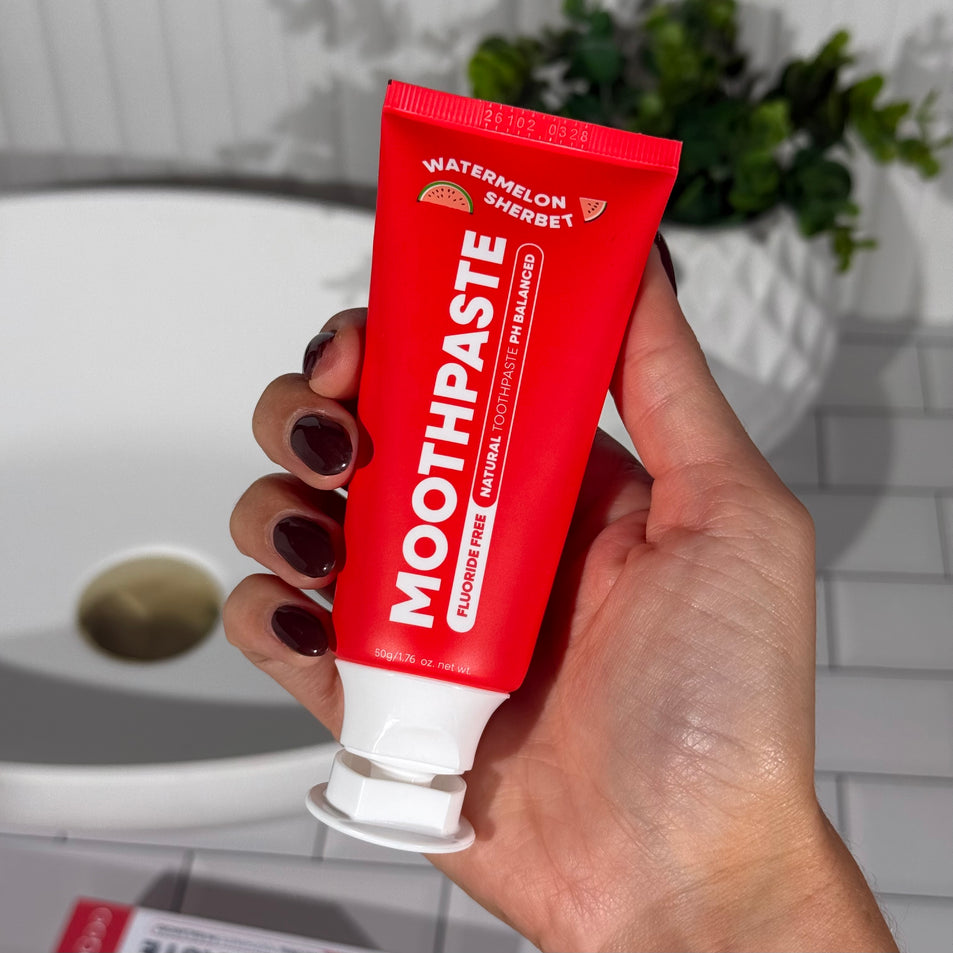 Hand holding MooGoo Mini Moo Kids Watermelon Sherbet flavoured toothpaste, with greenery and white tiles in the background. Healthy habit building, tastes like watermelon. Fluoride-free formula, safe for kids. Safe if swallowed.