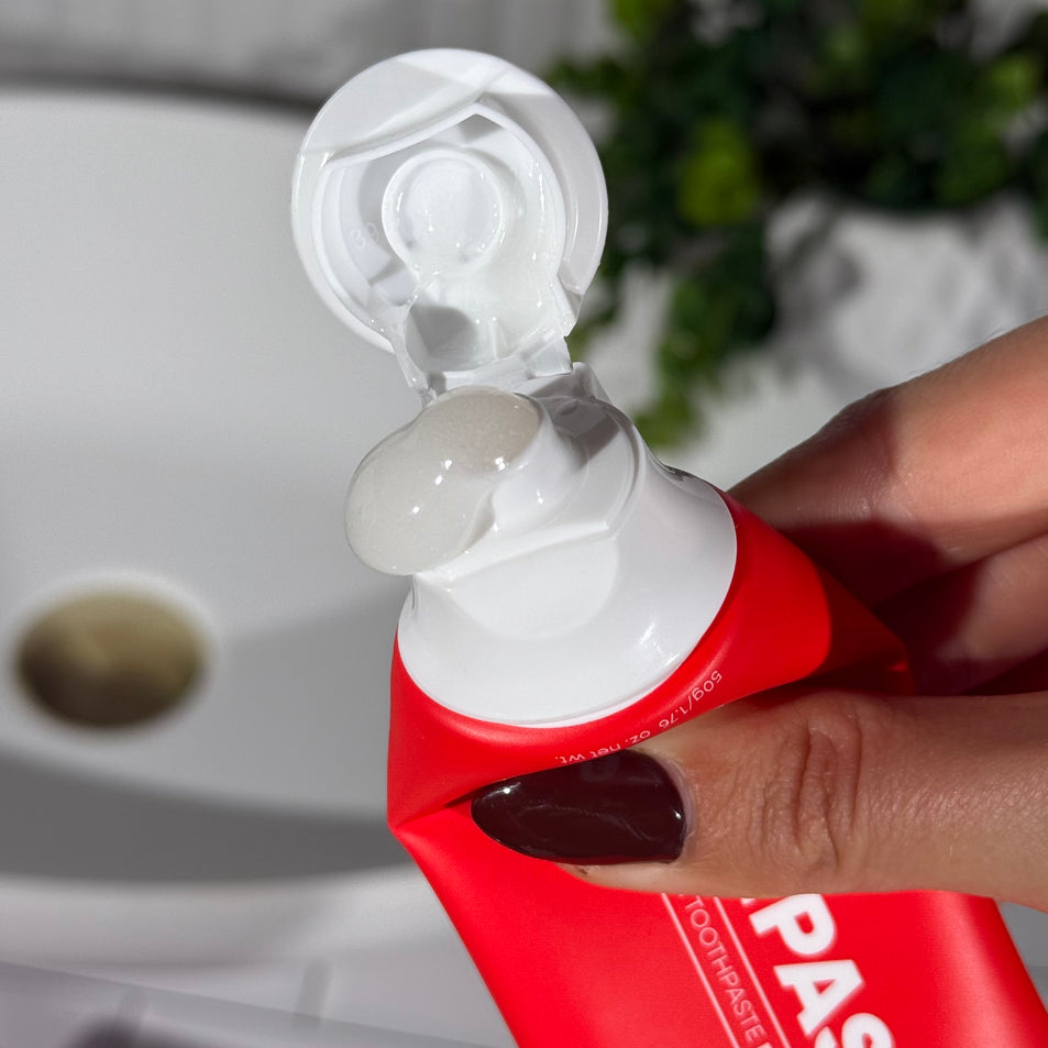 Hand holding a tube of MooGoo Mini Moo Watermelon Sherbet kids toothpaste and squeezing out the clear toothpaste. Healthy habit building, tastes like watermelon. Fluoride-free formula, safe for kids. Safe if swallowed. 