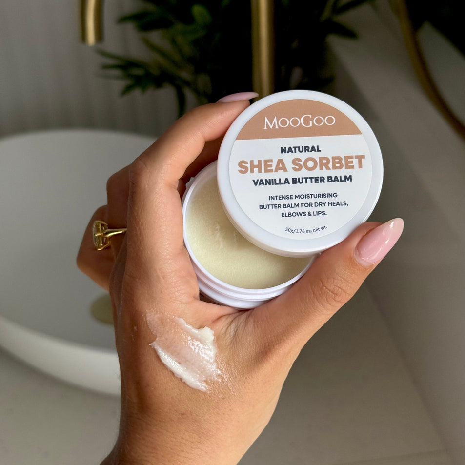 A hand holding an open jar of MooGoo Natural Shea Sorbet Vanilla Butter Balm, with some white balm smeared on the back of the hand, set against a bathroom sink background. Natural butter balm for dry, chapped and cracked skin. With Hemp, Shea and Oils.
