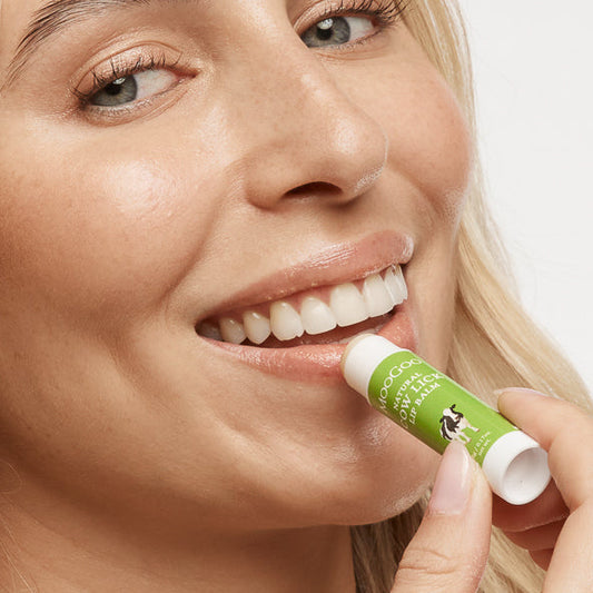 Woman applying MooGoo Cow Lick Lip Balm on plain background, natural lip moisturiser with edible ingredients for smooth soft lips, petroleum free, all natural.